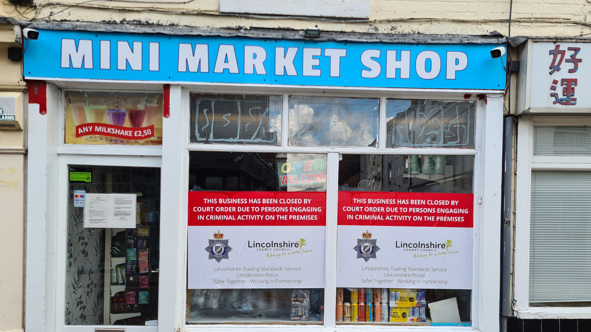 A small convenience shop in a town. A pale blue banner at the top of the shop reads Mini Market Shop