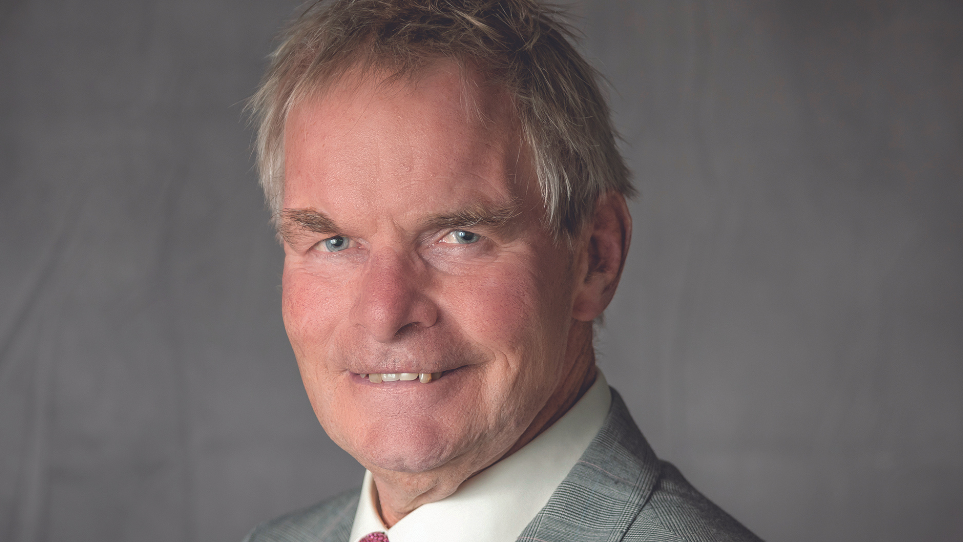 A man sat smiling to the camera in a head shot with a grey back ground. The man is dressed smart in a suit and tie.