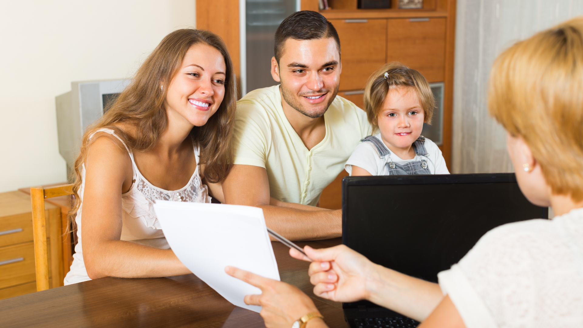 A family sat at a desk with a lady sat in front of a laptop opposite them. The lady sat near the laptop is holding a pen and pointing to a piece of paper.