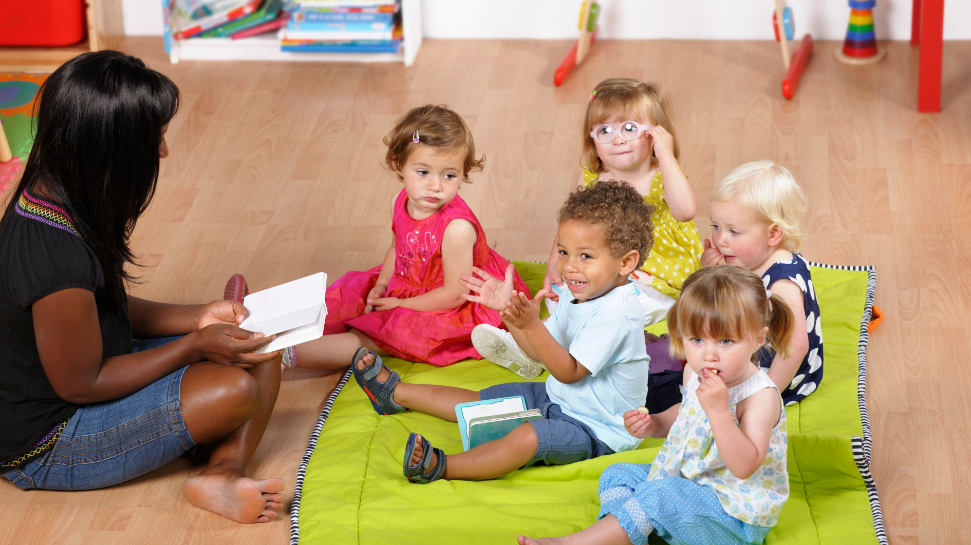 Childminder sat on the floor reading five children a story, who are sat on a green matt.