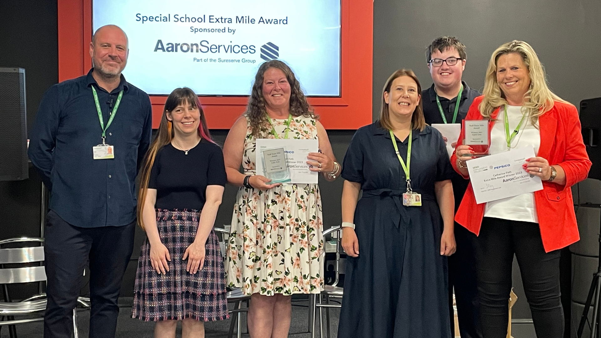 Six professionals stood up wearing lanyards in smart casual wear in front of a presentation for an awards ceremony