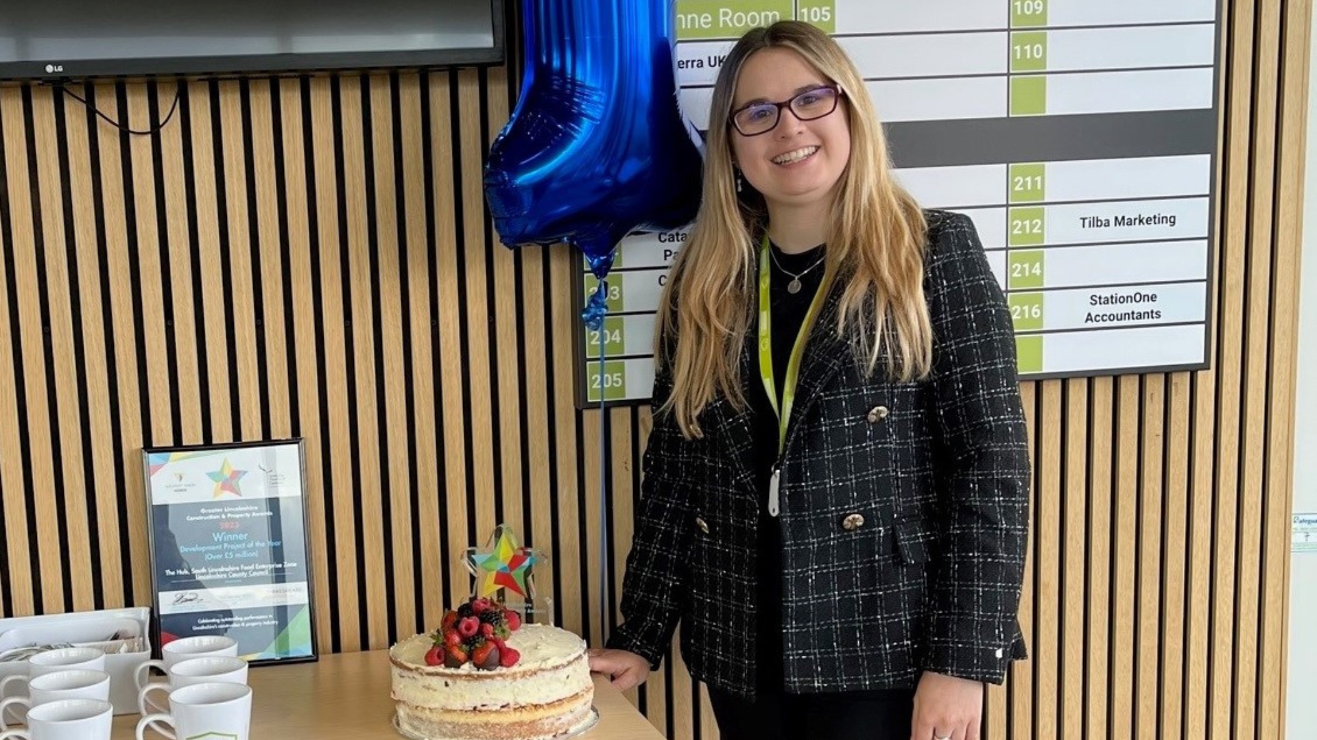 A young lady with blonde hair smiles standing next to a birthday cake