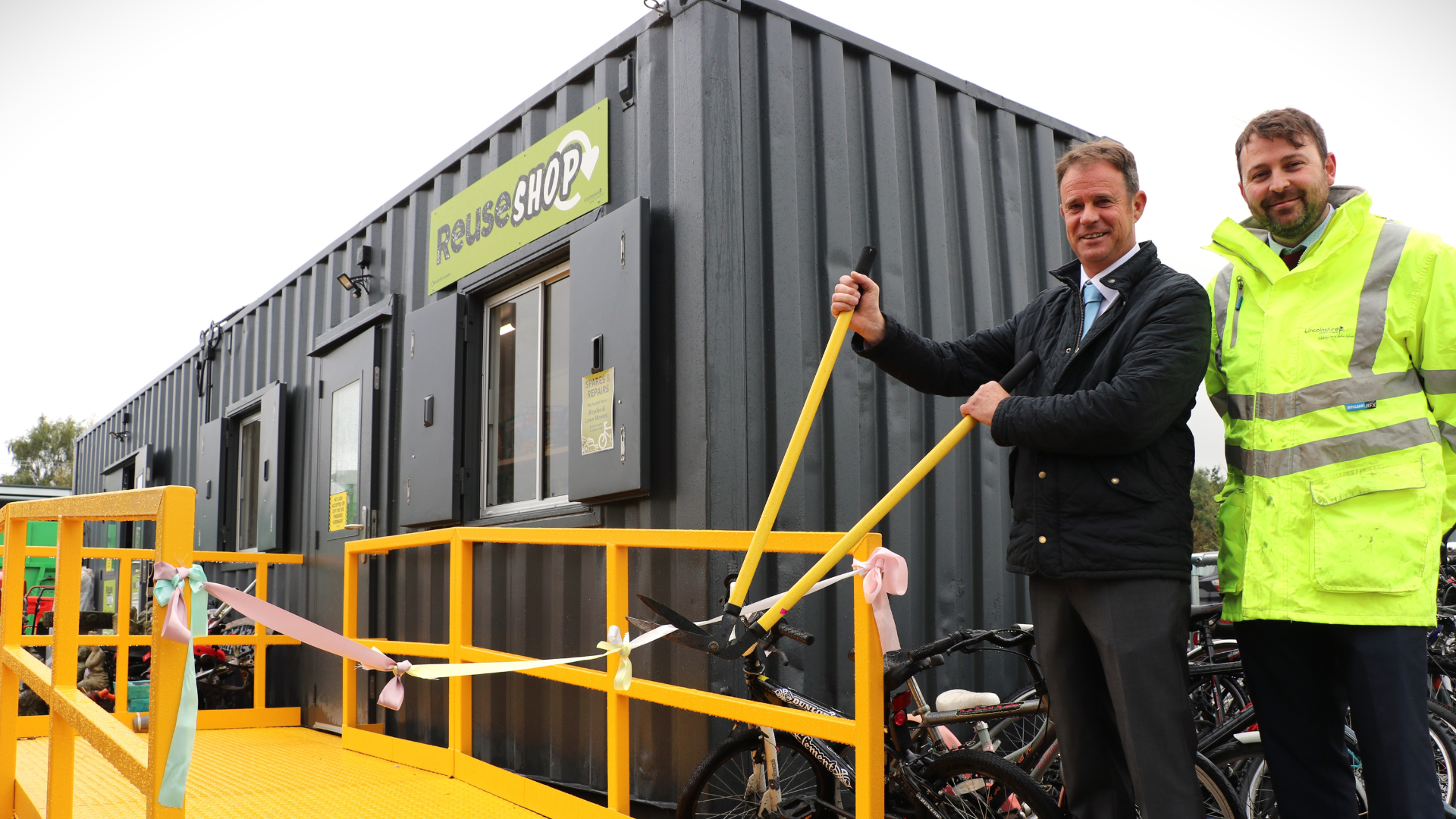 Two men stand in front of a recycled shipping container. One is cutting a ribbon with a pair of garden shears, the other is wearing a high-vis jacket.